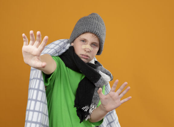 小生病的小男孩戴着暖和的帽子 戴着围巾 裹着毛毯 鼻子上贴着一块补丁 他看起来很不高兴 很不舒服 双手站在橙色的墙上做着防御的手势穿制造手