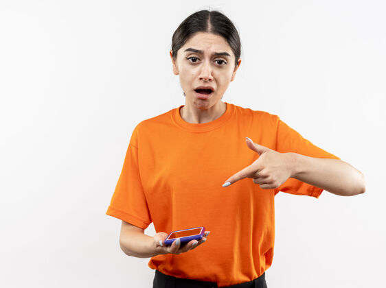 女孩穿着橙色t恤的年轻女孩拿着智能手机 用食指指着它 站在白色的墙上感到困惑和不快指着手指困惑