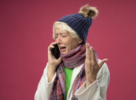 帽子不舒服的生病的不健康的女人 短头发 戴着暖和的围巾和帽子 感觉不舒服 一边用手机说话一边高举手臂站在粉红色的墙上哭泣生病手机不健康