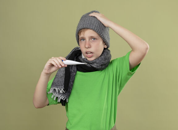 困惑生病的小男孩穿着绿色t恤 戴着暖和的围巾和帽子 感觉不舒服 拿着温度计 看起来很困惑 站在光墙上的温度光温暖温度