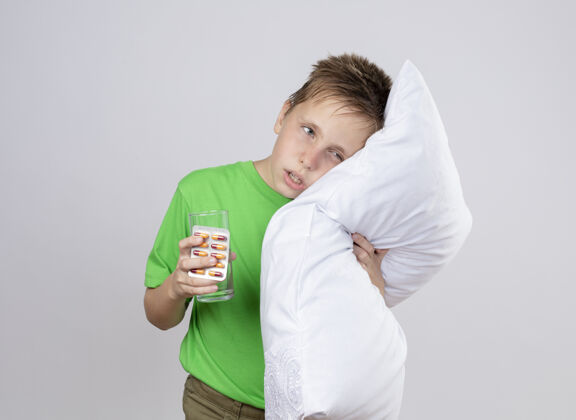 玻璃生病的小男孩穿着绿色t恤感到不舒服抱着枕头和一杯水靠在枕头上站在白色的墙上站着头不舒服