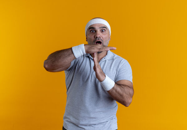 姿势戴着头巾的成熟的运动型男人望着前面 双手站在橙色的墙上 做着暂停的手势头带时间教练