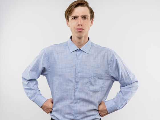 臀部身穿蓝色衬衫的年轻人 表情严肃自信 双臂放在臀部 站在白墙上站立表情自信