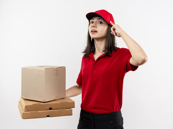 女孩年轻的送货女孩穿着红色制服 戴着帽子 拿着盒子和披萨盒 用食指指着太阳穴 站在白色的墙上 看上去自信地专注于一项任务帽子专注太阳穴