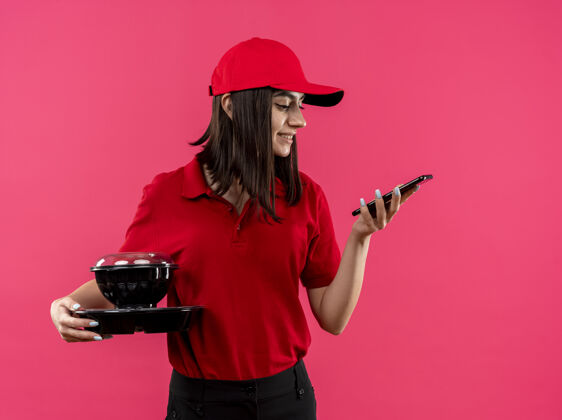 送货穿着红色马球衫 戴着帽子 拿着食品包的年轻送货女孩站在粉红色的墙上微笑着看着她的智能手机屏幕食品马球年轻