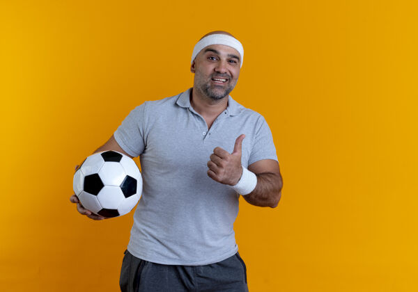 看戴着头巾的成熟运动型男子举着足球望着前方 脸上带着微笑 竖起大拇指站在橙色的墙上肌肉运动足球