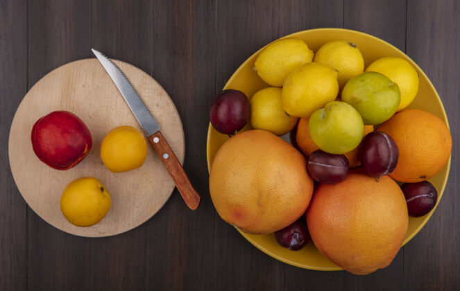 杏子俯瞰黄色碗里的柠檬 橘子 李子和葡萄柚 还有杏子和桃子 架子上放着一把刀 背景是木制的柠檬李子木头