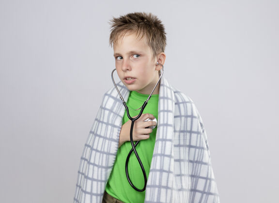 毯子生病的小男孩穿着绿色t恤 裹着毛毯 脖子上戴着听诊器 站在白色的墙上 听着心跳混乱包裹感觉站