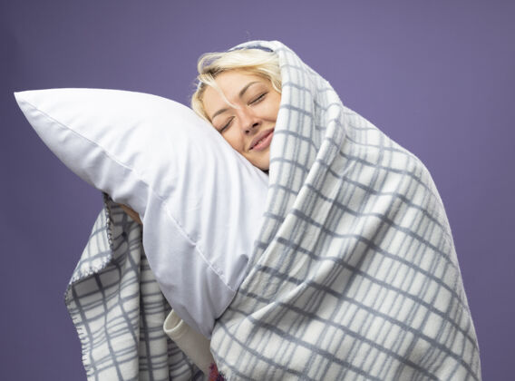帽子生病的不健康的女人 短发 戴着暖和的帽子 裹着毯子 抱着枕头 把头靠在枕头上 闭着眼睛 在紫色的墙上微笑头毯子关闭
