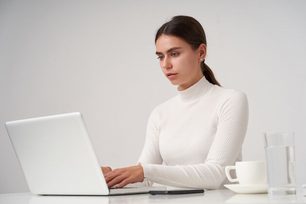 穿着严肃的年轻漂亮的黑发女性坐在白色墙壁上的桌子旁 凝神地看着屏幕 一边用键盘打字键盘Mua站着
