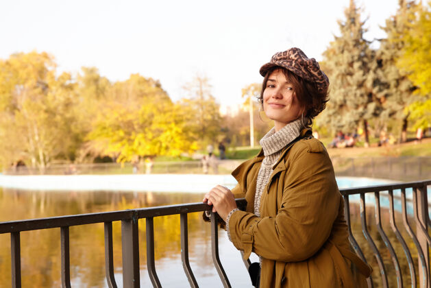 女性积极的年轻迷人的短发黑发女性斜靠在铁栏杆上 在城市公园的湖边摆姿势 愉快地看着 温柔地微笑着树欢呼休闲