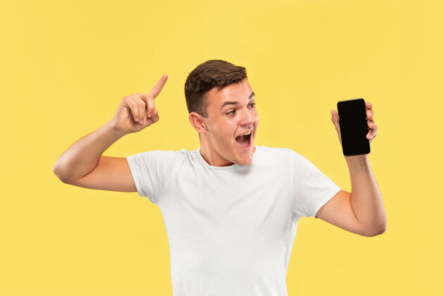 成功黄色工作室背景上的白人年轻人半身肖像穿着衬衫的漂亮男模人类情感的概念 面部表情 销售 广告展示手机屏幕和微笑商人工作员工