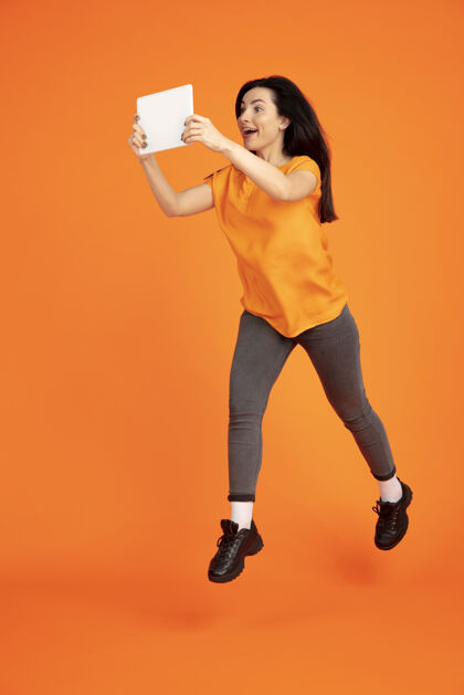 行政橙色工作室背景上的白种人年轻女子肖像穿着衬衫的漂亮深色女性模特人类情感的概念 面部表情 销售 广告广告空间平板电脑跳跃财务衬衫看起来