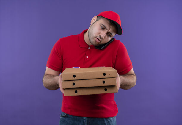 困惑站在紫色的墙上 一个身穿红色制服 戴着帽子 拿着披萨盒 拿着手机的年轻送货员看起来很困惑站着送货市民