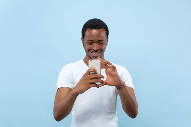 胡须在蓝色背景上 一个身穿白衬衫的非洲裔美国年轻人的半身特写肖像人类的情感 面部表情 广告概念在他的智能手机上拍照或视频博客内容企业家长相工作