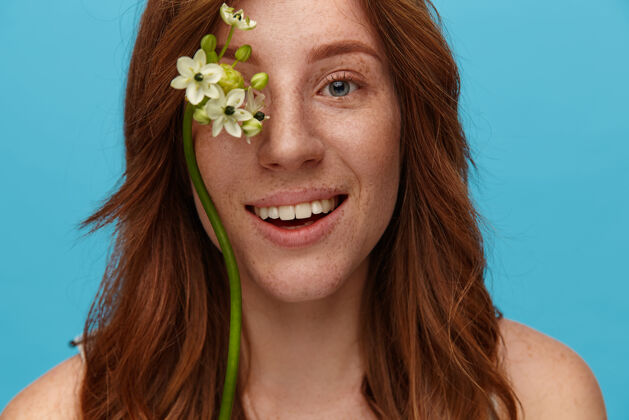 肩膀迷人的年轻红发女性的肖像 波浪式的发型 脸上挂着鲜花 对着镜头开心地微笑 在蓝色的背景下与世隔绝Swoosh表情花