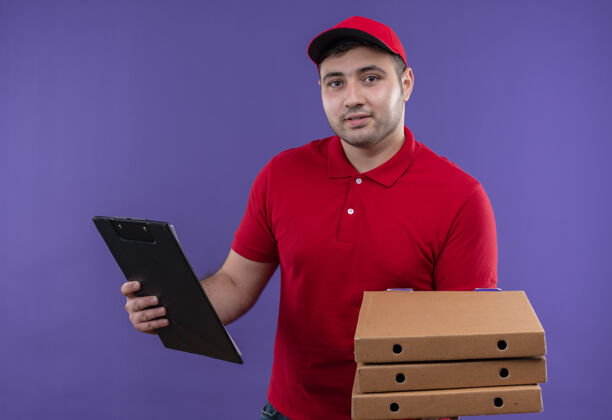披萨身穿红色制服 头戴鸭舌帽 手持披萨盒和剪贴板的年轻送货员站在紫色的墙上 脸上洋溢着幸福的笑容人男人人