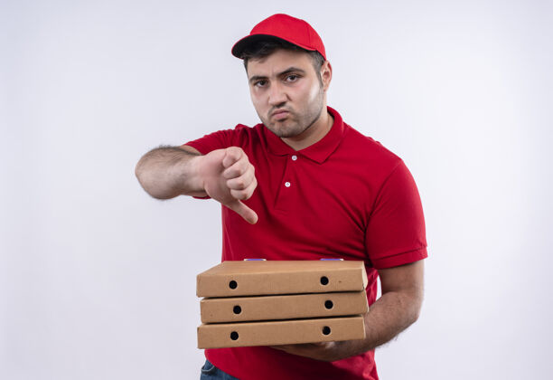 送货年轻的送货员穿着红色制服 戴着帽子 手里拿着比萨饼盒 不高兴地竖起大拇指站在白色的墙上年轻人帽子人