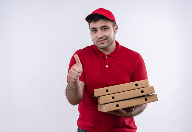 制服年轻的送货员身穿红色制服 头戴鸭舌帽 手里拿着披萨盒 站在白色的墙边高兴地竖起大拇指男人人欢呼