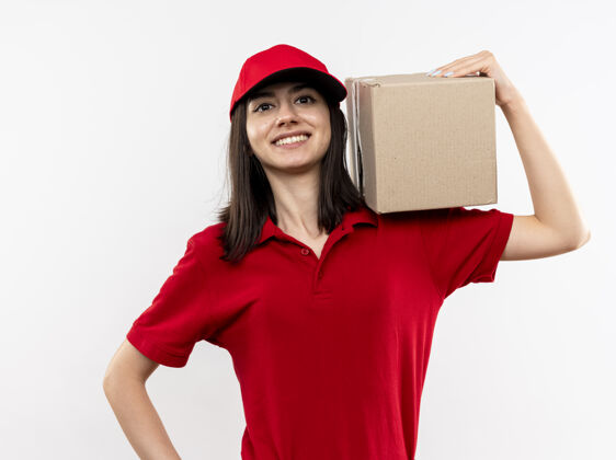 年轻年轻的送货员穿着红色制服 戴着帽子 拿着盒子 站在白色的背景下 脸上带着大大的笑容 看起来很自信举行脸帽子