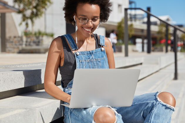 耳机照片中的黑人高兴的女人浏览网页 键盘上的笔记本电脑的反馈或评论 听在线广播耳机 穿着破烂的工作服 做远程工作 模型外冲浪遥控台阶