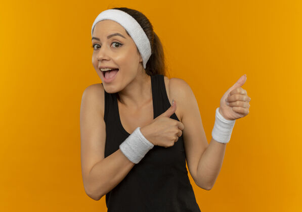 手指身穿运动服的年轻健身女士戴着头巾 高兴而又惊讶地用手指着站在橙色墙上的一边运动女人运动装