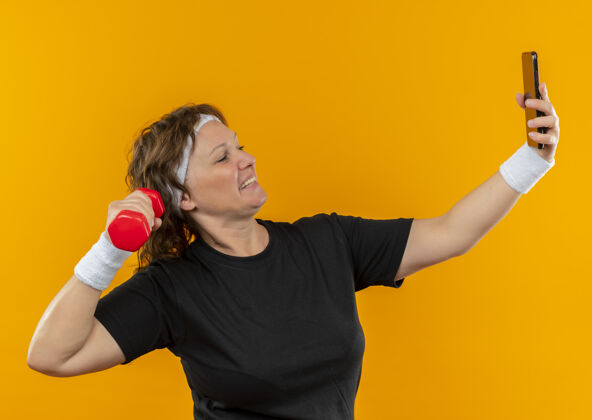 头带身穿黑色t恤 头箍 手持智能手机哑铃的中年运动女性站在橙色的墙上自拍人运动中间