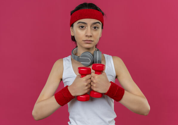运动身穿运动服 头箍 举着两个哑铃 表情严肃地站在粉色墙壁上锻炼身体的年轻健身女士人运动员人