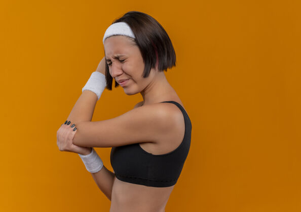 看穿着运动服的年轻健身女 头箍看起来不舒服 她站在橘色的墙上摸着她的胳膊肘感到疼痛头带运动疼痛