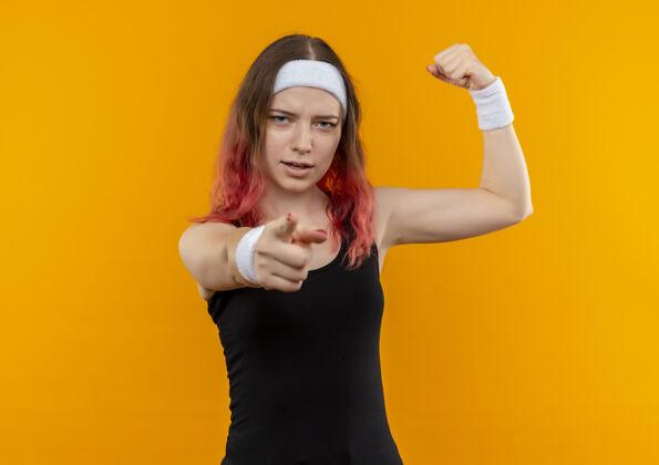 运动身着运动服的年轻健身女士举起拳头 自信地用食指指着站在橙色墙上的摄像机手指运动健身