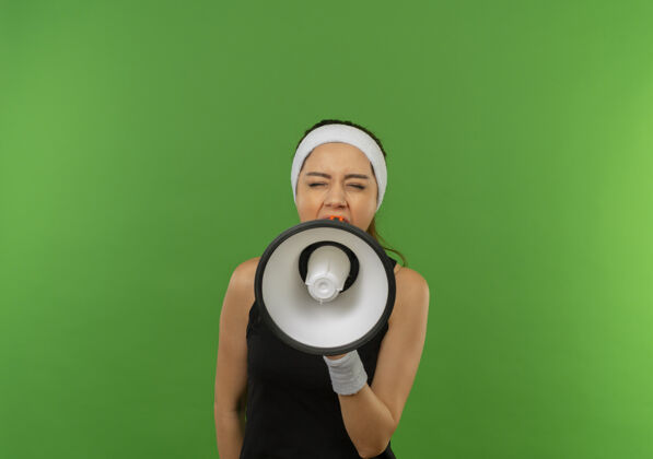 健身戴着头巾的年轻健身女士站在绿色的墙上对着扩音器大声喊叫 表情咄咄逼人女市民人