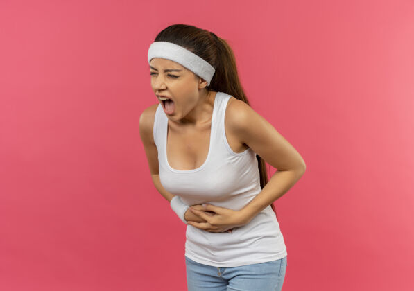 胃痛穿着运动服的年轻健身女士戴着头巾 看起来不舒服 摸着肚子 站在粉红色的墙上胃疼不适姿势人