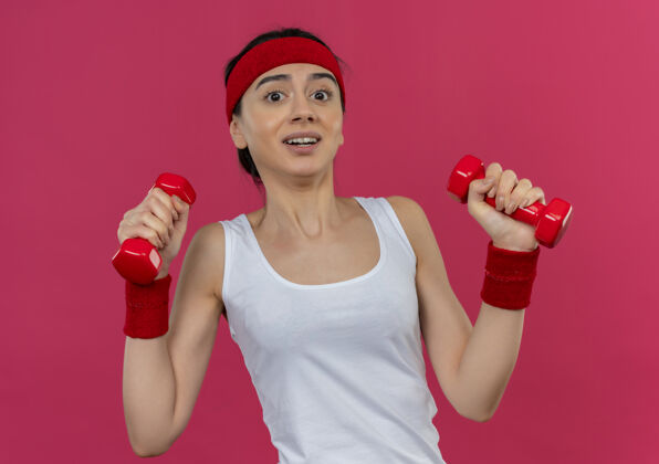 惊人身穿运动服的年轻健身女士 头戴钢带 双手举着两个哑铃做运动运动装人站着