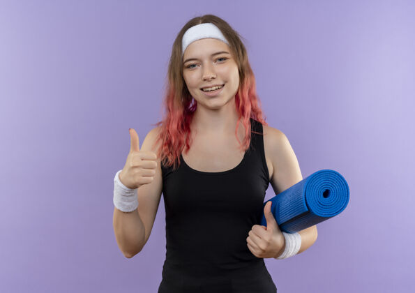 运动身穿运动服 手持瑜伽垫 微笑着 竖起大拇指的年轻健身女士站在紫色的墙上运动员抱着站着