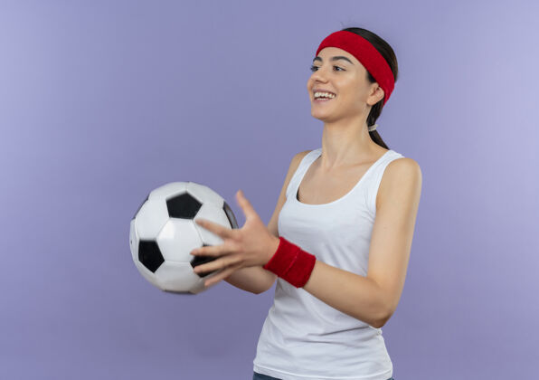 运动身穿运动服 头箍 手持足球 微笑 自信 快乐 积极向上的年轻健身女士站在紫色的墙上站立人年轻