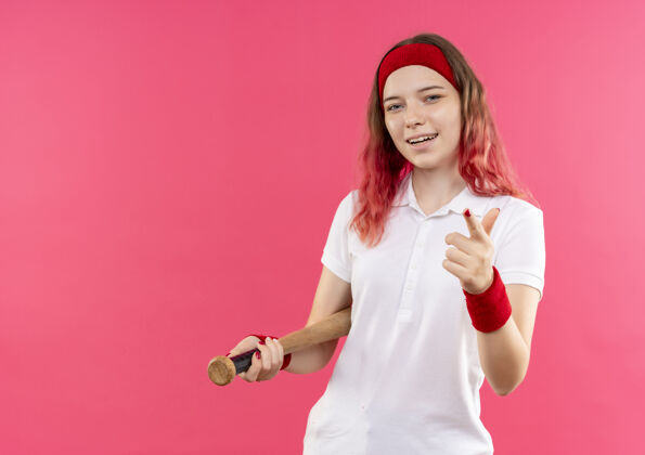 运动装戴着头巾的年轻运动女性手持球棒 手指指着镜头 站在粉色的墙上 开心地微笑着站着微笑欢呼