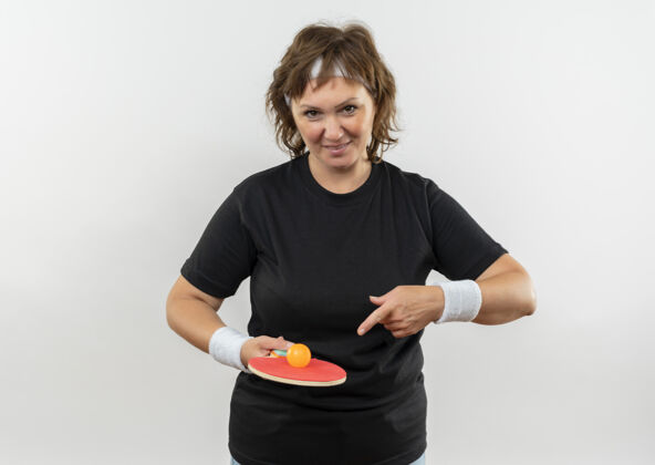 运动中年运动型女子 身穿黑色t恤 头戴头巾 手持乒乓球球拍 手指指着球拍 站在白墙上 面带笑容快乐微笑运动装