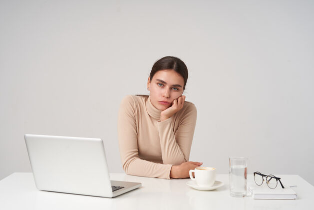 优雅无聊的年轻迷人的黑发女性坐在桌子旁 头靠在举起的手上 在办公室里用笔记本电脑工作 看着的时候保持嘴唇合拢心情头发手