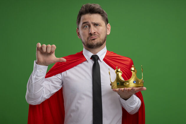 皇冠身穿红色斗篷的超级英雄商人手持王冠 站在绿色背景上 指着它困惑超级英雄指着拿着