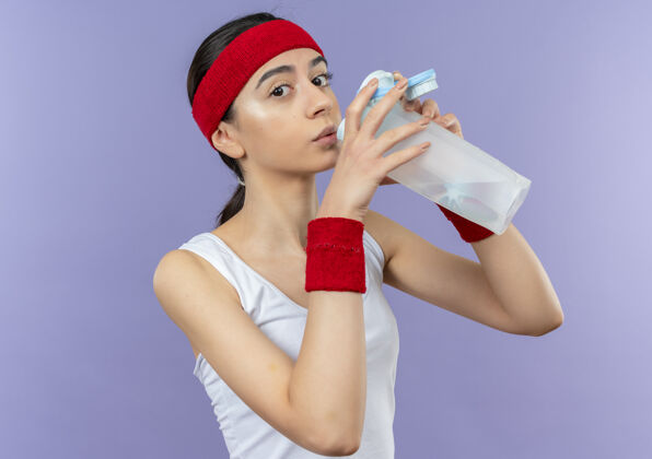 健身穿着运动服的年轻健身女士戴着头巾站在紫色的墙上喝水 看起来很累水站市民