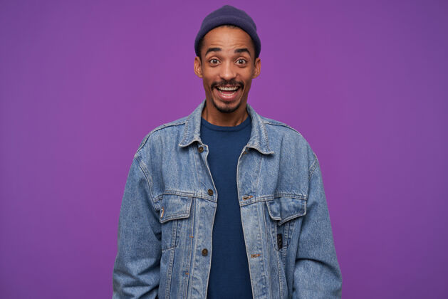 毛衣室内镜头：年轻快乐的黑皮肤男性 留着胡须 表情激动 眉头高抬 笑容满面 在紫色的墙上摆姿势胡须积极情绪