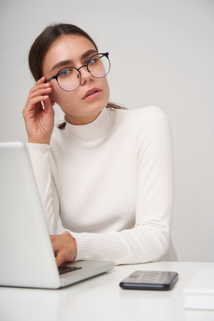 工作一张室内照片 年轻漂亮的黑发女子戴着眼镜 正坐在白色墙壁上的桌子旁 手里拿着一台现代化的笔记本电脑 身着正装 神情严肃桌子20多岁女士