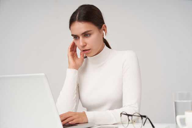 情绪专注年轻漂亮的黑发女性戴着耳机坐在白色的墙上 在办公室工作的时候专注地看着笔记本电脑的屏幕年轻女性欧式