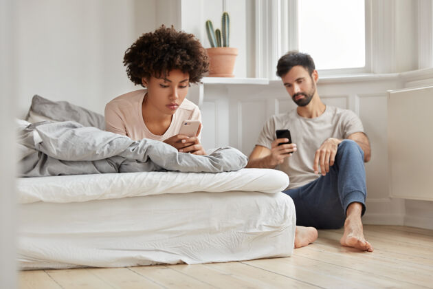 聊天多民族夫妻通过社交网络观看视频 手持现代电影 妻子躺在床上 丈夫坐在地板旁 写短信 在家里宽敞明亮的房间里摆姿势在线交流设备男人短信