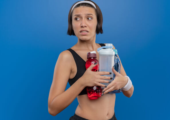 困惑穿着运动服的年轻健身女士拿着两瓶水站在蓝色的墙上 困惑地看着一边运动装人瓶子