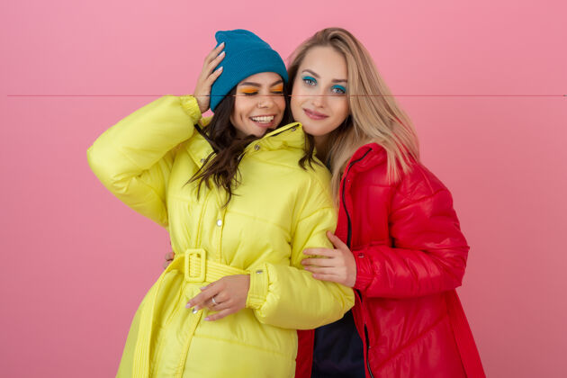 颜色两位迷人的活跃女性穿着鲜艳的红黄相间的冬季羽绒服在粉色的墙上摆着姿势 朋友们在一起玩得很开心 温暖的外套时尚潮流 疯狂搞笑的面孔羽绒服冬天外观