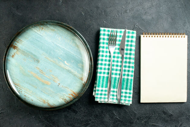 培养皿俯视图圆形拼盘餐刀和叉子在绿色和白色方格餐巾笔记本在黑色的桌子上刀顶部玻璃