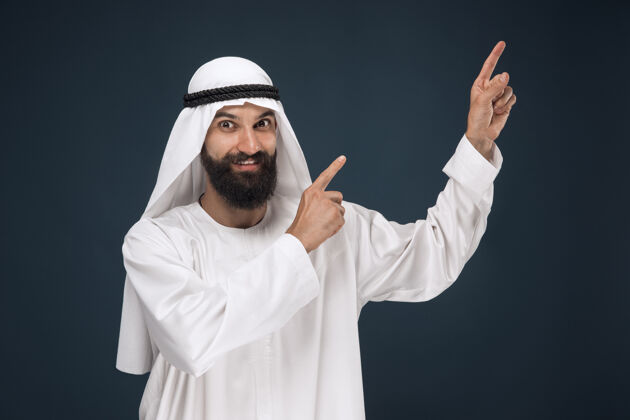 胡须深蓝色工作室背景上的半身阿拉伯沙特商人肖像年轻男模微笑着指着或选择商业概念 金融 面部表情 人类情感男人脸惊喜
