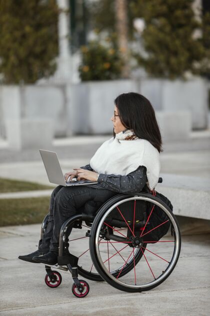 垂直坐在轮椅上拿着笔记本电脑的女人的侧视图虚弱户外残障