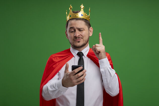手机穿着红色斗篷戴着王冠的超级英雄商人用智能手机显示食指微笑 站在绿色背景上有新想法目录手指创意